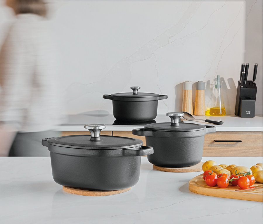 Drei schwarze Kochtöpfe mit Deckel auf einer Küchentheke, daneben ein Schneidebrett mit Tomaten und Kartoffeln.