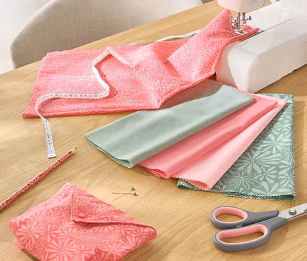 Auf einem Tisch liegen ein 5-teiliges Stoff-Set, eine Nähmaschine, eine Schere, ein Bleistift und ein Maßband.