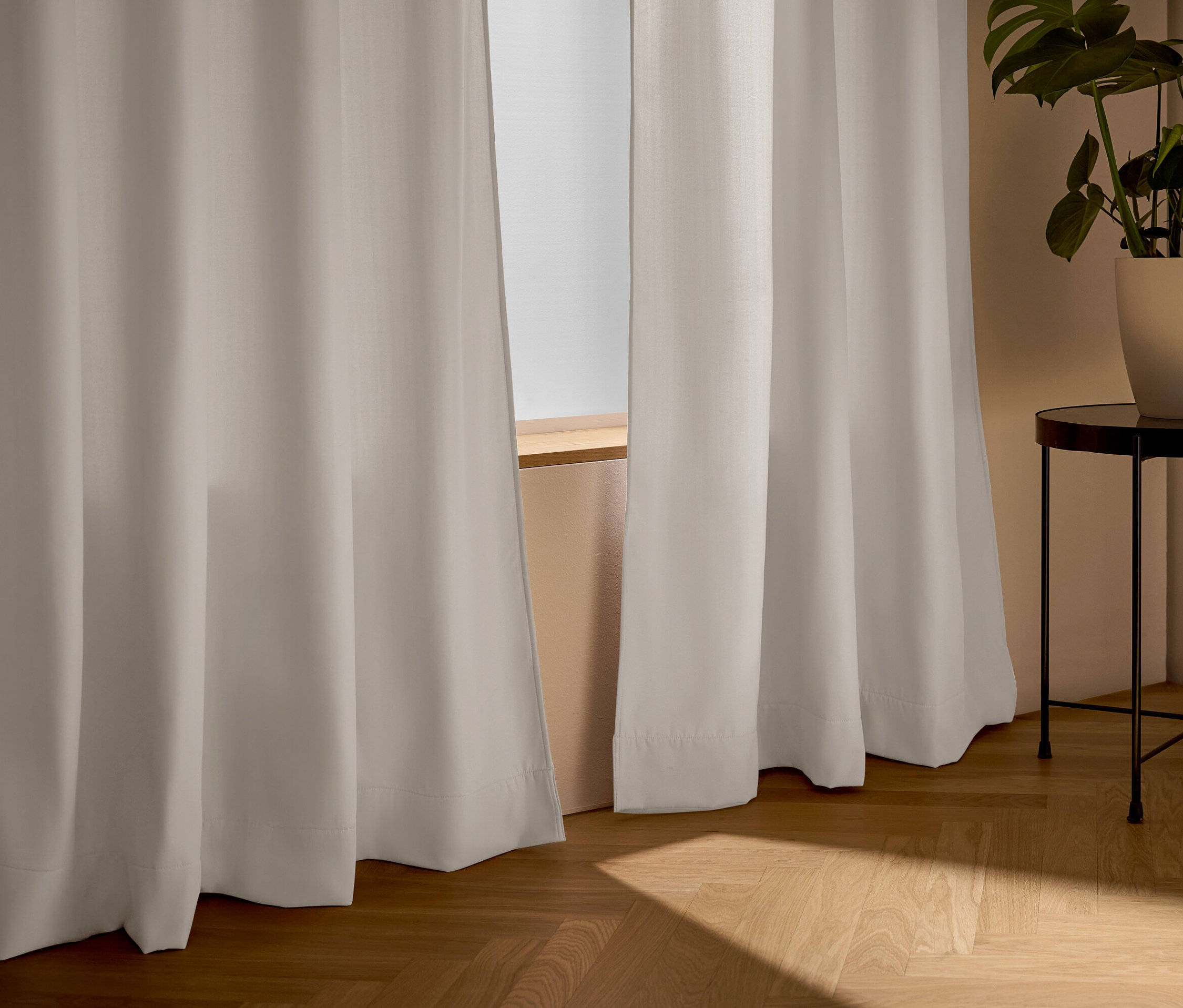 Beige Vorhänge vor einem Fenster, daneben ein Tisch mit einer Pflanze.