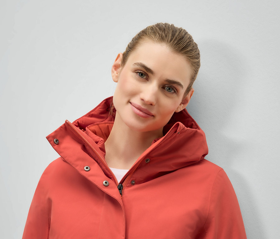 Porträt einer Frau in einer korallfarbenen Jacke mit Kapuze.
