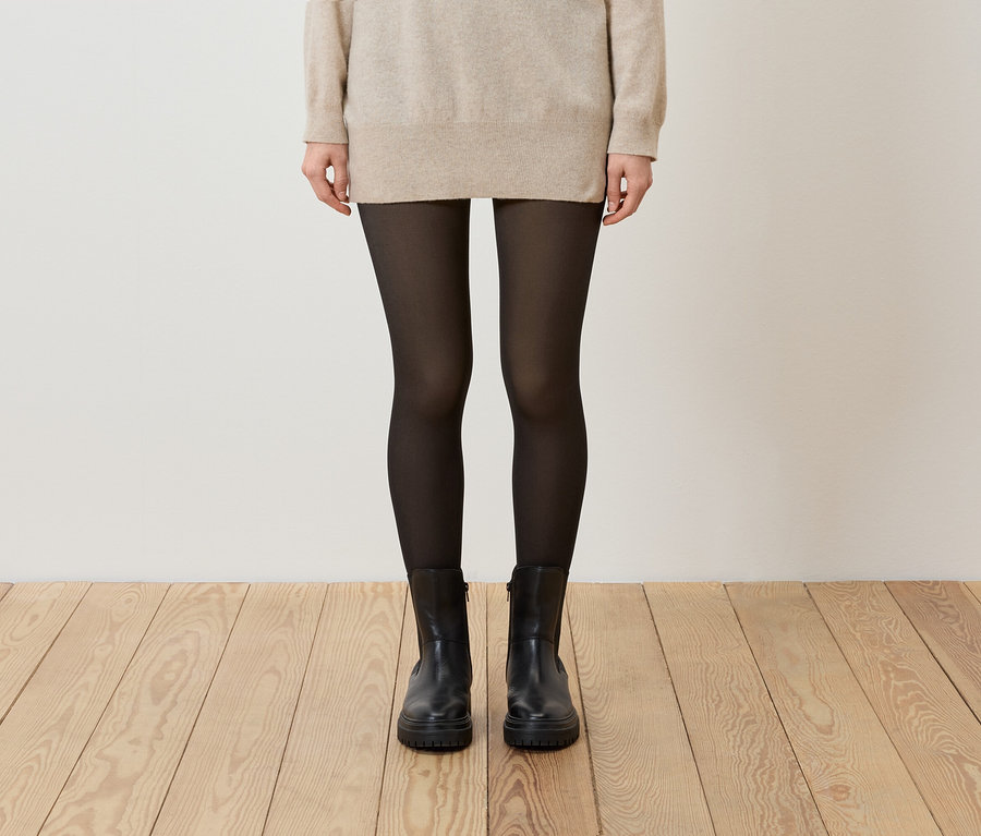 Frau trägt beigen Pullover, schwarze Strumpfhose und schwarze Stiefel.