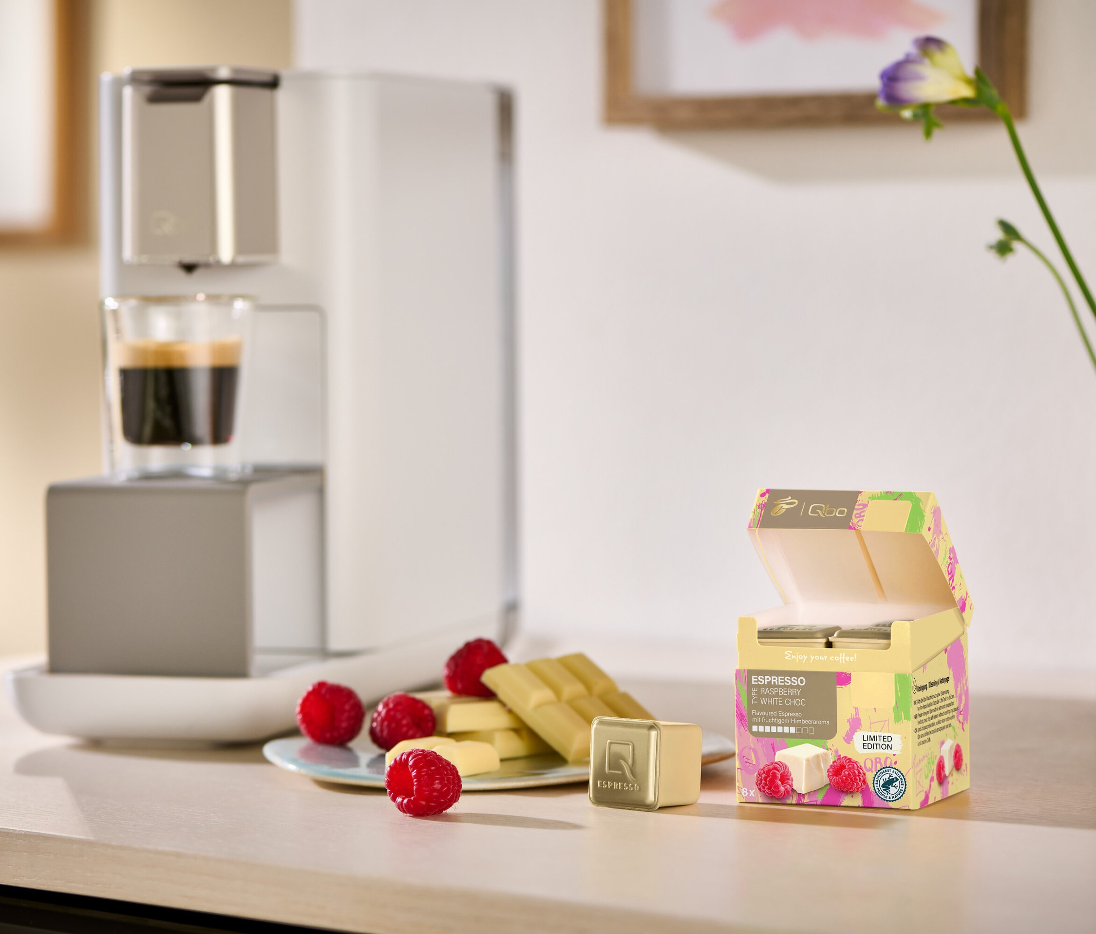 Espresso und weiße Schokolade mit Himbeeren auf einem Tisch neben einer Kaffeemaschine. Im Hintergrund eine ISCC RA Qbo ES Rasp. White Choc 8 Cup.