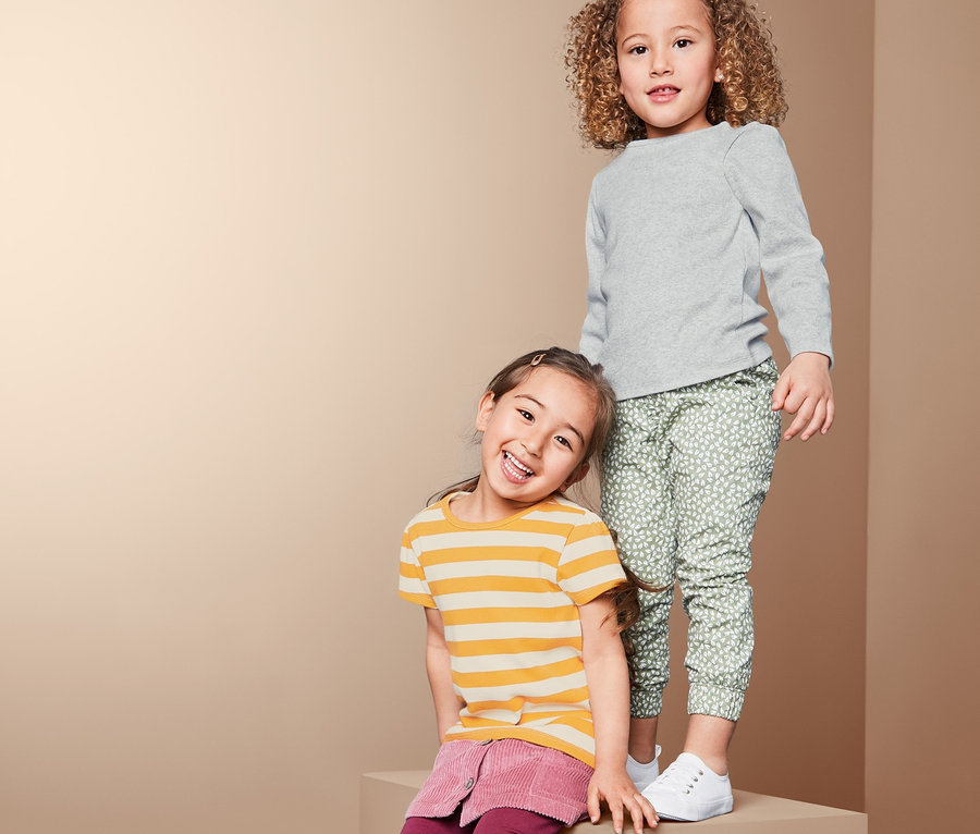 Zwei Mädchen posieren auf einem Block. Eines trägt ein graues Rippenshirt und eine grüne Hose. Das andere trägt ein gelb gestreiftes Shirt und einen bordeauxroten Rock.