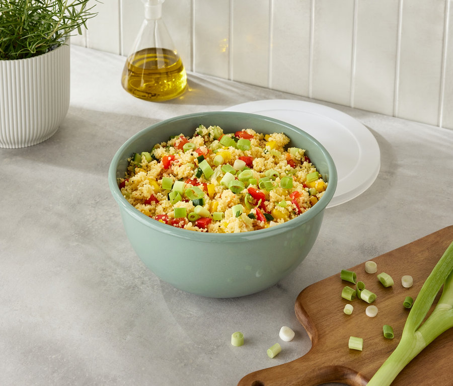 Eine grüne Schüssel mit Couscous und Gemüse neben Zwiebeln auf einem Schneidebrett.