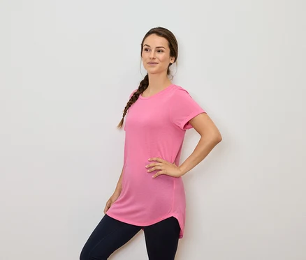 Frau mit braunen Haaren im Zopf posiert in einem rosa T-Shirt und dunkelblauen Leggings.