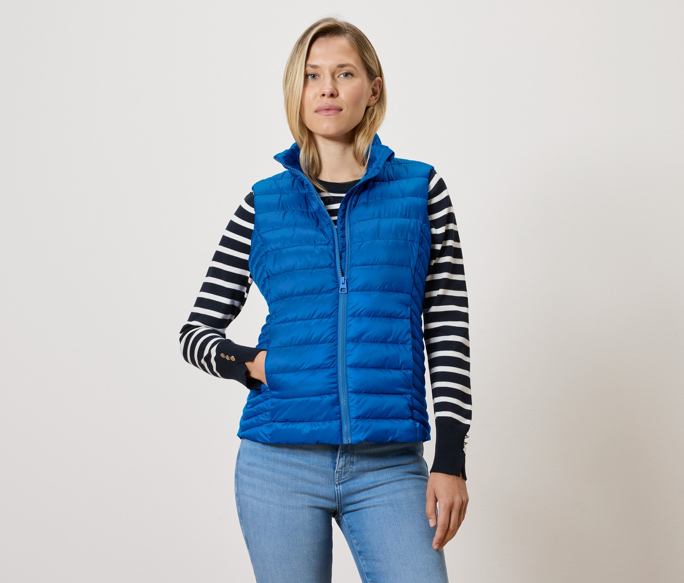 Frau trägt eine blaue Steppweste mit Reißverschluss, einen schwarz-weiß gestreiften Pullover und blaue Jeans.