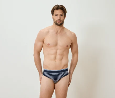 Mann mit freiem Oberkörper und gestreifter Unterhose steht vor einer weißen Wand.