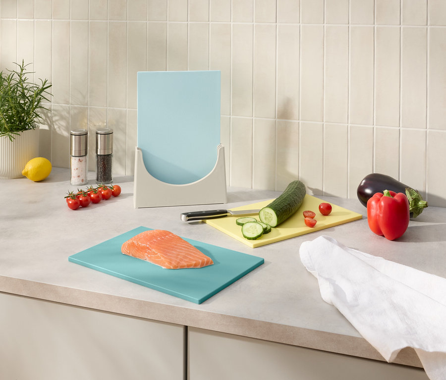 Auf einer Küchentheke stehen ein Topf mit Rosmarin, Salz- und Pfefferstreuer, ein Zitronen, eine blaue Schneidebrettablage mit einem blauen Schneidebrett, ein Messer und verschiedene Gemüsesorten wie eine Aubergine und eine Paprika.