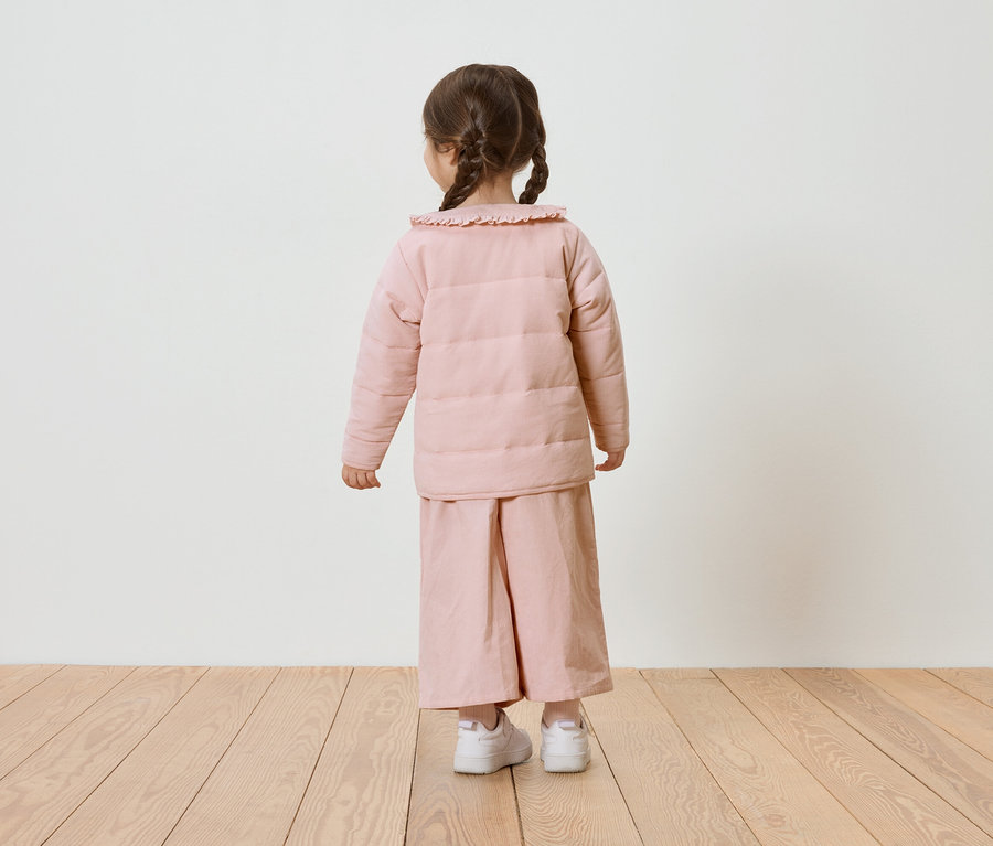Kind mit Zöpfen, von hinten gesehen, trägt eine rosa Pufferjacke und weite Hose, dazu weiße Turnschuhe.