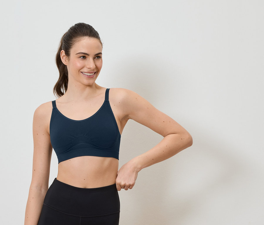Lächelnde Frau mit Pferdeschwanz trägt einen marineblauen Sport-BH und schwarze Leggings vor einem weißen Hintergrund.
