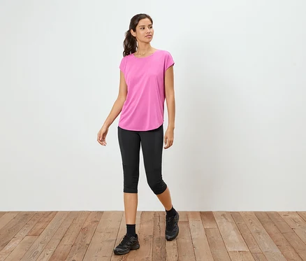 Frau steht auf Holzboden und trägt ein pinkes T-Shirt, schwarze Capri-Leggings und schwarze Turnschuhe.