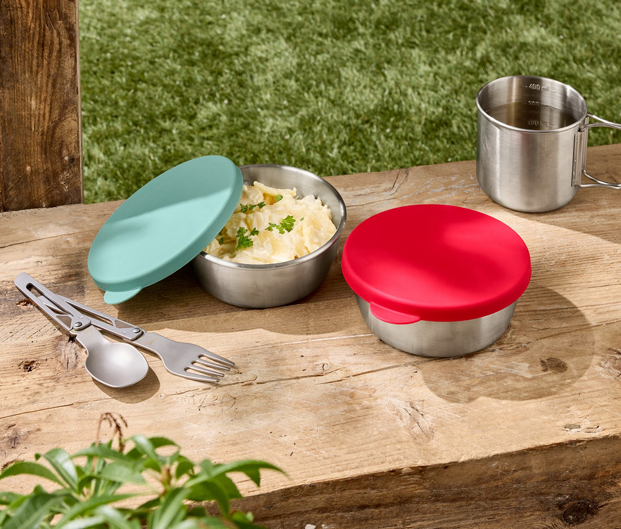 Zwei Edelstahlschüsseln mit Deckel, eine davon mit Kartoffelsalat, daneben ein silberner Becher und ein Besteckset auf einem Holztisch.
