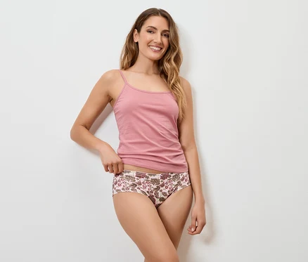 Eine Frau mit langen, gewellten, blonden Haaren lächelt und posiert in einem rosafarbenen Tanktop und einer floralen Unterhose vor einer weißen Wand.