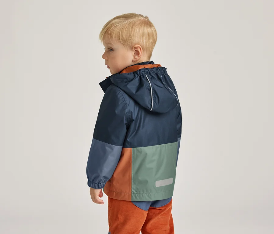 Kleiner Junge mit blonder Kurzhaarfrisur, von hinten gesehen, trägt eine farblich abgesetzte Regenjacke in Blau, Grün und Orange.