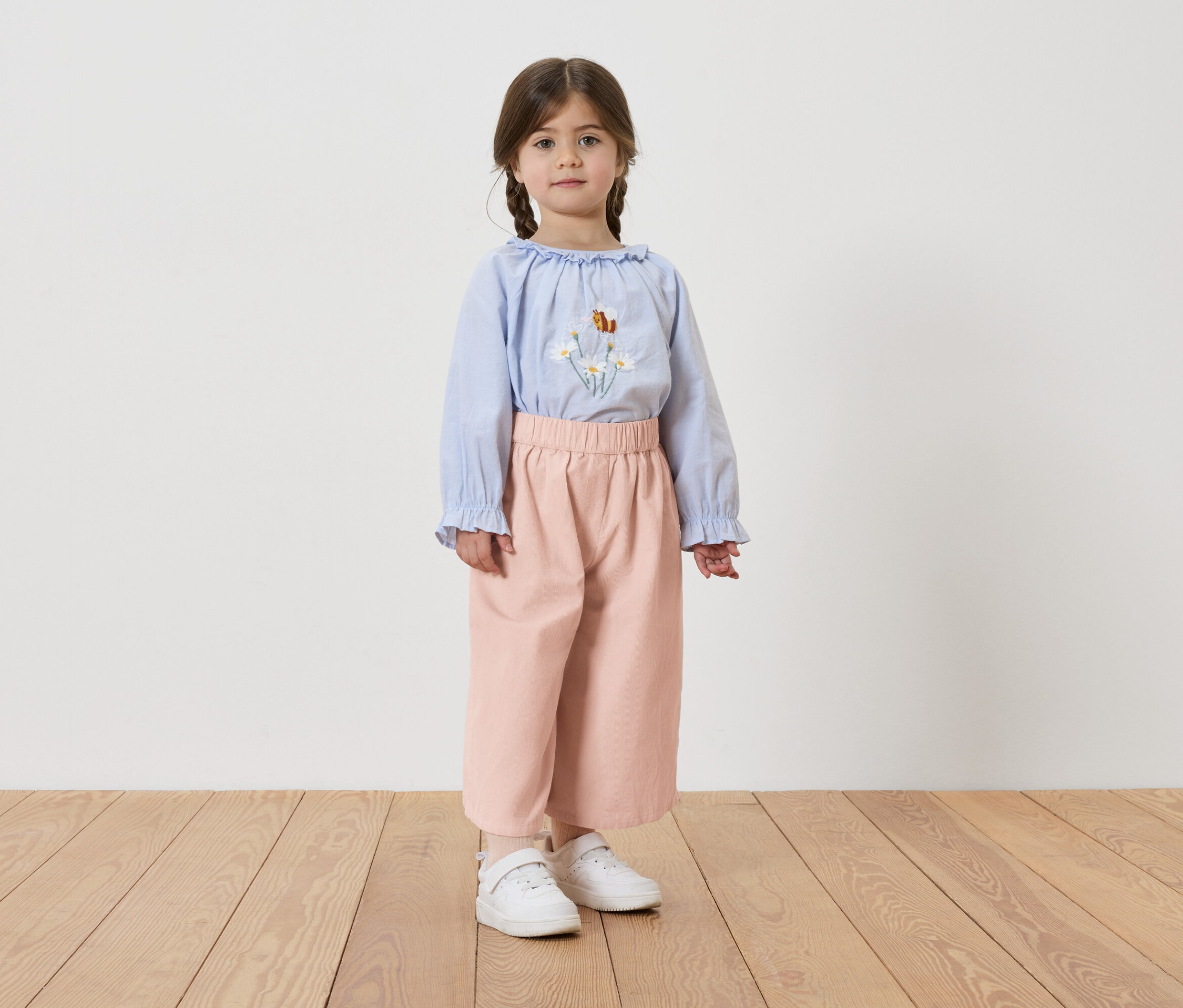 Mädchen mit Zöpfen, trägt eine hellblaue Bluse mit Blumenstickerei, rosafarbene Hose und weiße Turnschuhe.