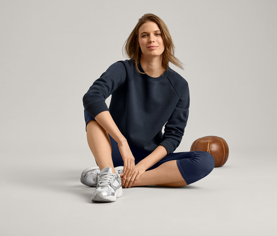 Frau sitzt im Schneidersitz, trägt ein marineblaues Sweatshirt, marineblaue Leggings und silberne Turnschuhe.