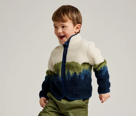 Ein kleiner Junge mit braunen Haaren, der eine Fleecejacke in Weiß, Grün und Blau sowie grüne Hosen trägt, lächelt und blickt nach rechts.