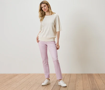 Eine Frau steht mit einem cremefarbenen Strickpullover, einer lila Hose und weißen Turnschuhen vor einer weißen Wand.