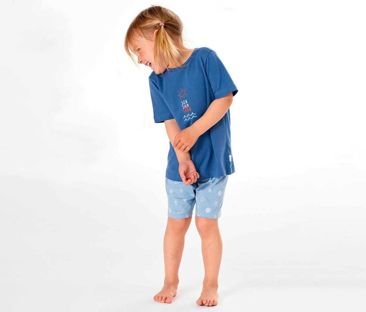 Mädchen trägt SCHIESSER kurzen Kinder-Pyjama mit blauem T-Shirt und gemusterten Shorts, lächelnd zur Seite.
