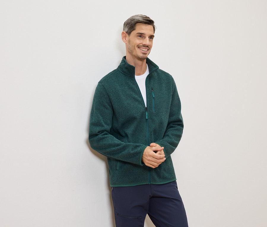 Mann mit grünem Fleecepullover und blauer Hose lehnt an einer weißen Wand und lächelt.