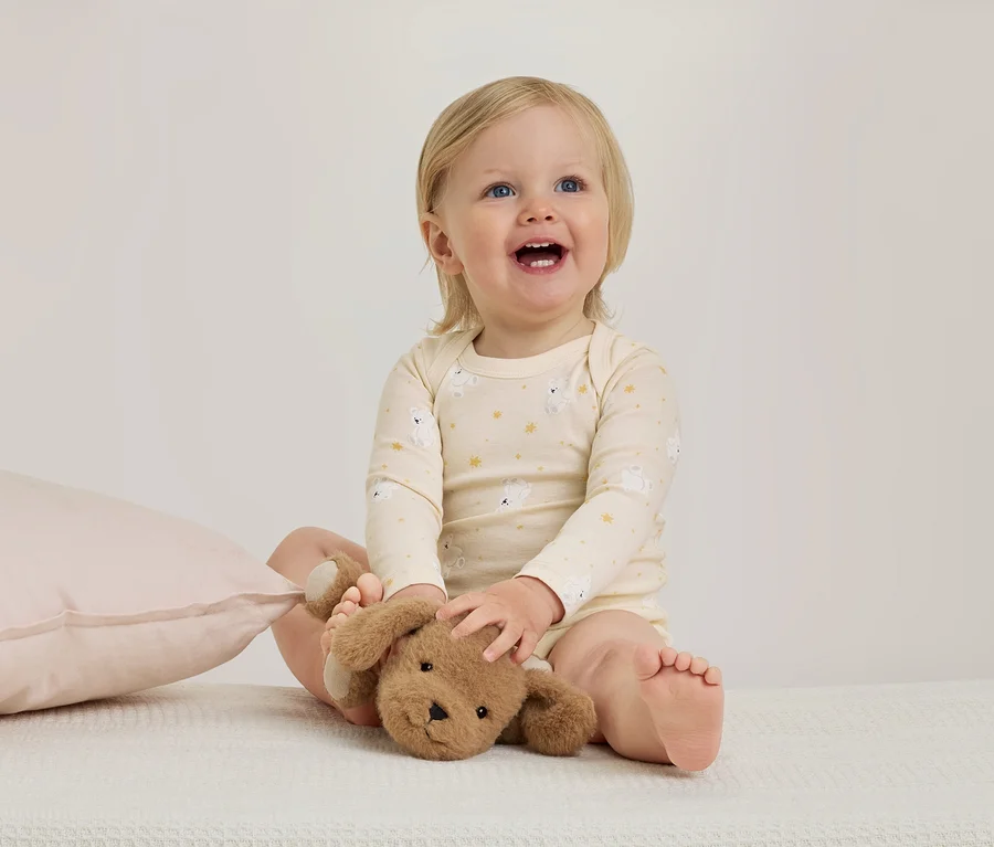 Ein lächelndes Baby mit blonden Haaren sitzt auf einer weißen Decke, trägt einen hellgelben Strampler mit Bärenmuster und hält ein braunes Plüschtier. Links liegt ein rosa Kissen.