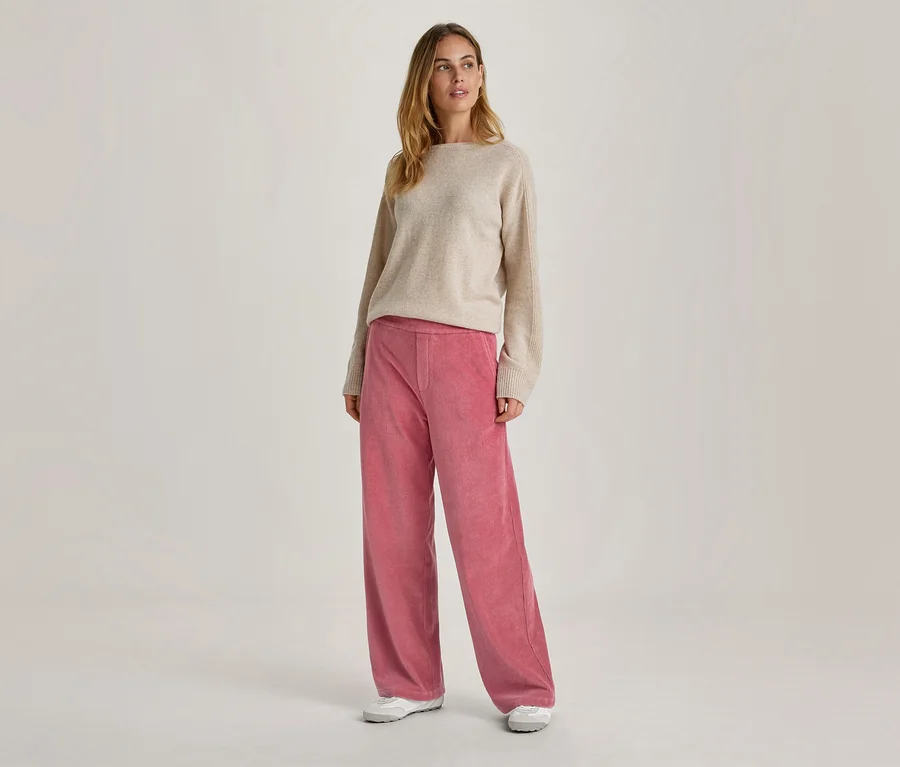 Eine Frau steht, nach rechts blickend, in einem beigen Pullover mit U-Boot-Ausschnitt, einer rosa weiten Hose und weißen Turnschuhen.