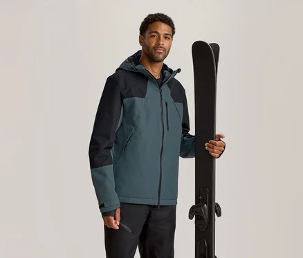 Mann in zweifarbiger petrolfarbener und schwarzer Skijacke, der schwarze Skier vertikal hält.
