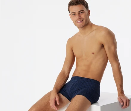 Oberkörperfreier Mann in dunkelblauen, karierten SCHIESSER Shorts, lächelnd auf einem weißen Würfel sitzend.