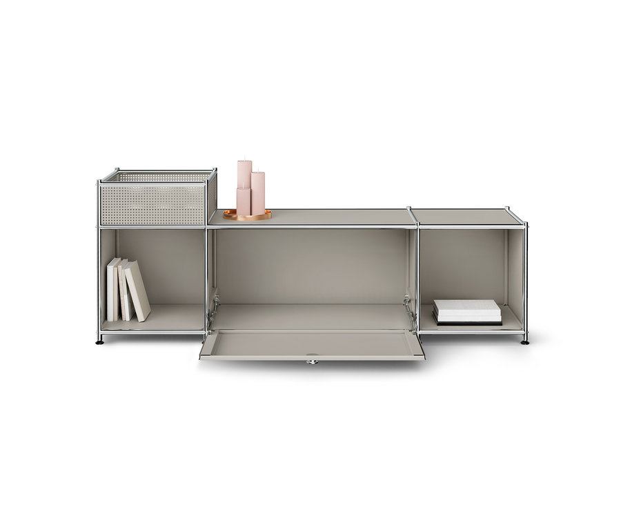 Hellgraues modulares Aufbewahrungsmöbel mit Chromrahmen. Der linke Bereich hat ein offenes Fach mit Büchern und einen perforierten Korb oben. Die Klapptür des mittleren Bereichs ist offen, und drei rosa Kerzen stehen auf seiner Oberfläche. Der rechte Bereich ist ein offenes Fach mit weißen Boxen.