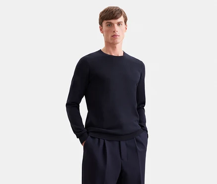 Mann in dunkelblauem Langarmpullover und dunkelblauer Hose, Hände in den Hosentaschen.