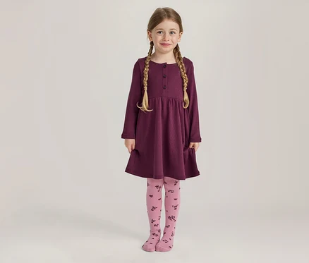 Ein junges Mädchen mit blonden Zöpfen lächelt stehend in einem langärmeligen pflaumenfarbenen Kleid mit Knöpfen und Taschen sowie rosa Strumpfhosen mit dunklem Blumenmuster.