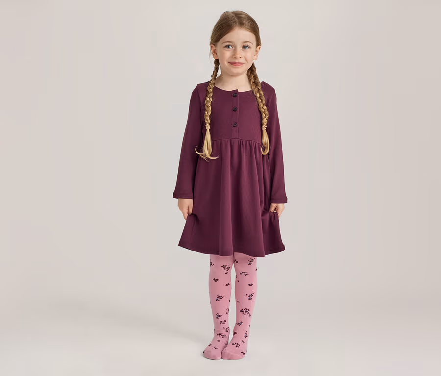 Ein junges Mädchen mit blonden Zöpfen lächelt stehend in einem langärmeligen pflaumenfarbenen Kleid mit Knöpfen und Taschen sowie rosa Strumpfhosen mit dunklem Blumenmuster.
