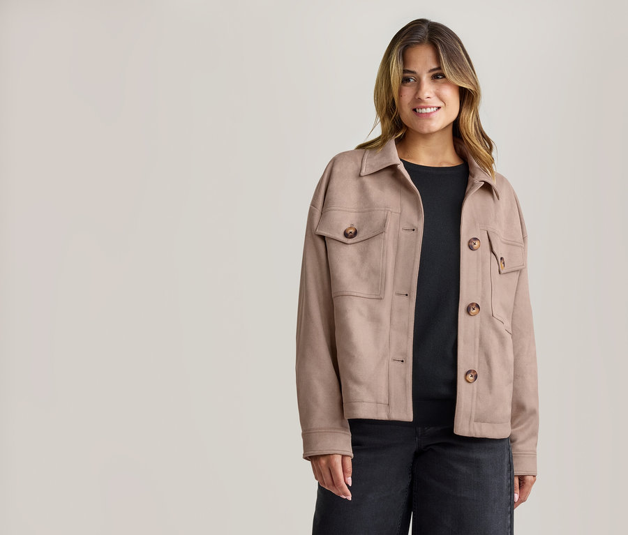 Lächelnde Frau in beiger Kragenjacke mit braunen Knöpfen und Taschen, schwarzem Oberteil und dunkler Jeans.
