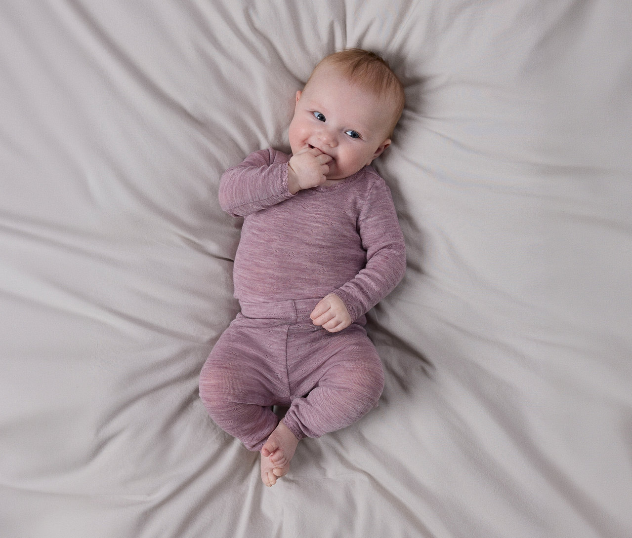 Lächelndes Baby liegt auf hellgrauem Laken, trägt langärmeliges, strukturiertes lila Oberteil und passende Hose, eine Hand am Mund.