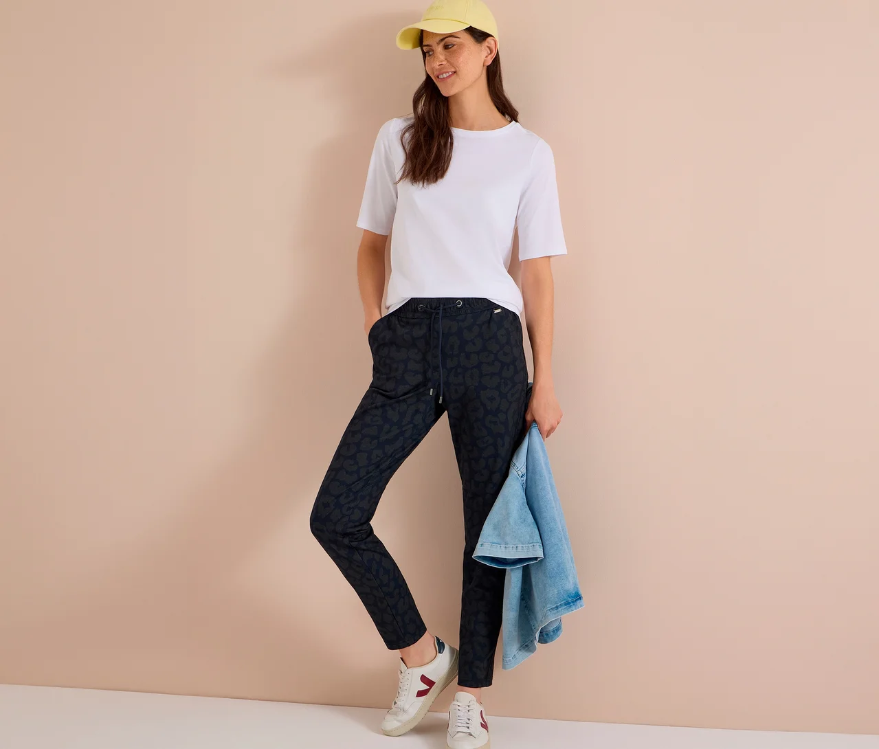 Frau trägt weißes T-Shirt, gelbe Kappe, weiße Sneaker und eine CECIL Jerseyhose mit Alloverprint, hält eine Jeansjacke.