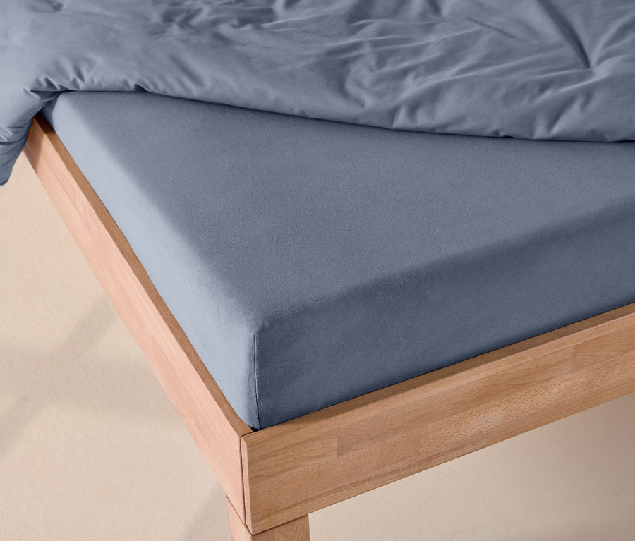 Bett mit blauem Jersey-Spannbettlaken und einer blauen Bettdecke auf einem hellen Holzrahmen.