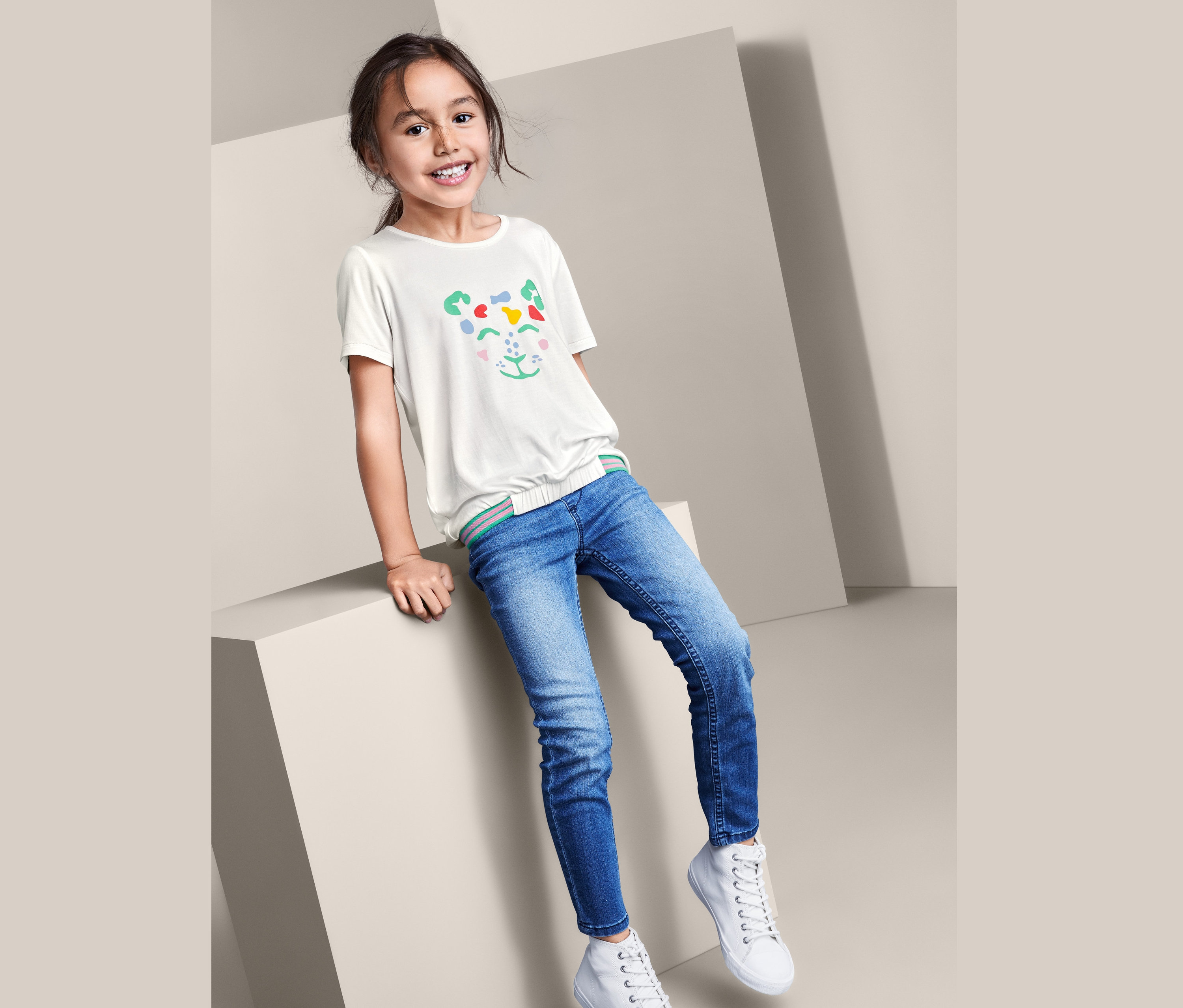 Ein Mädchen sitzt auf einem Würfel und trägt Jeans, weiße Turnschuhe und ein weißes T-Shirt in Schlupfform mit einem Tiergesicht.