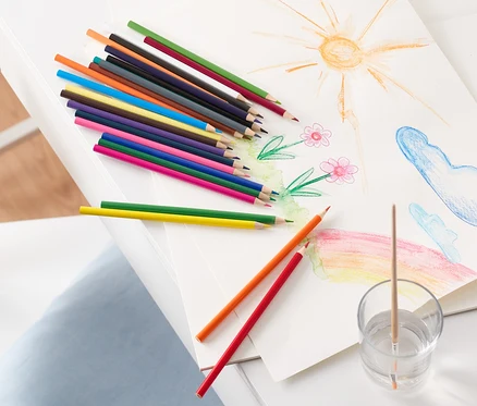 Auf einem weißen Tisch liegen eine Zeichnung von Sonne, Blumen, Wolken und einem Regenbogen, daneben Aquarell-Buntstifte und ein Glas mit Wasser und Pinsel.