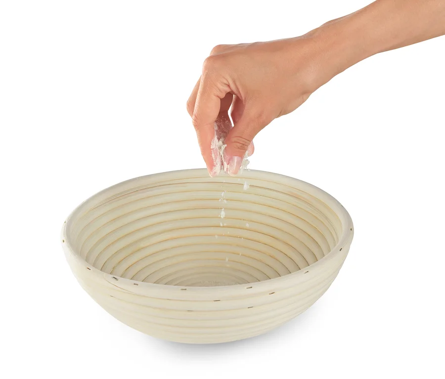 Eine Hand streut Mehl in ein rundes Gärkörbchen.