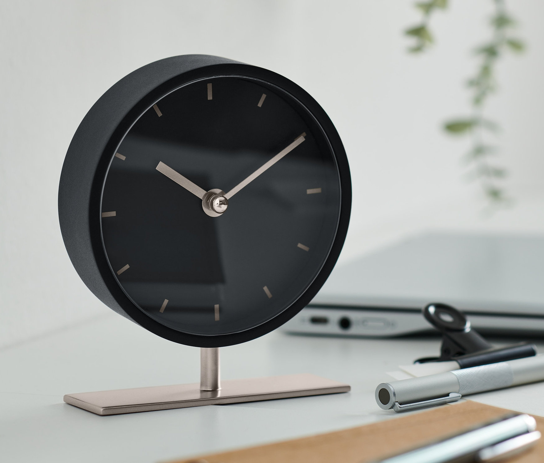 Eine schwarze Design-Tischuhr steht auf einem Tisch mit einem Laptop, einem Stift und einem Notizblock.