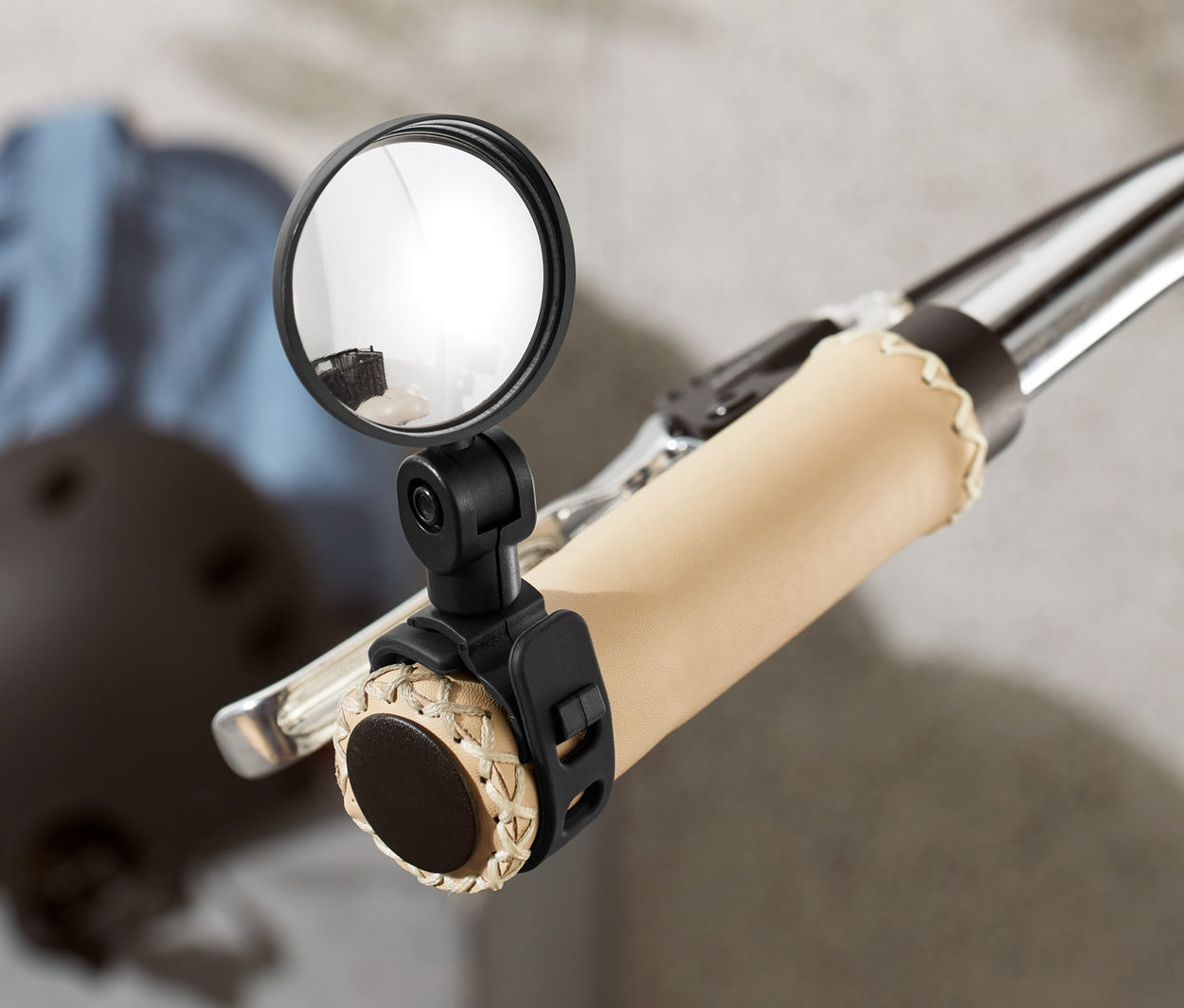 Nahaufnahme eines Fahrradlenkers mit montiertem Mini-Fahrradspiegel. Im Hintergrund sind ein Helm und eine Jacke zu sehen.