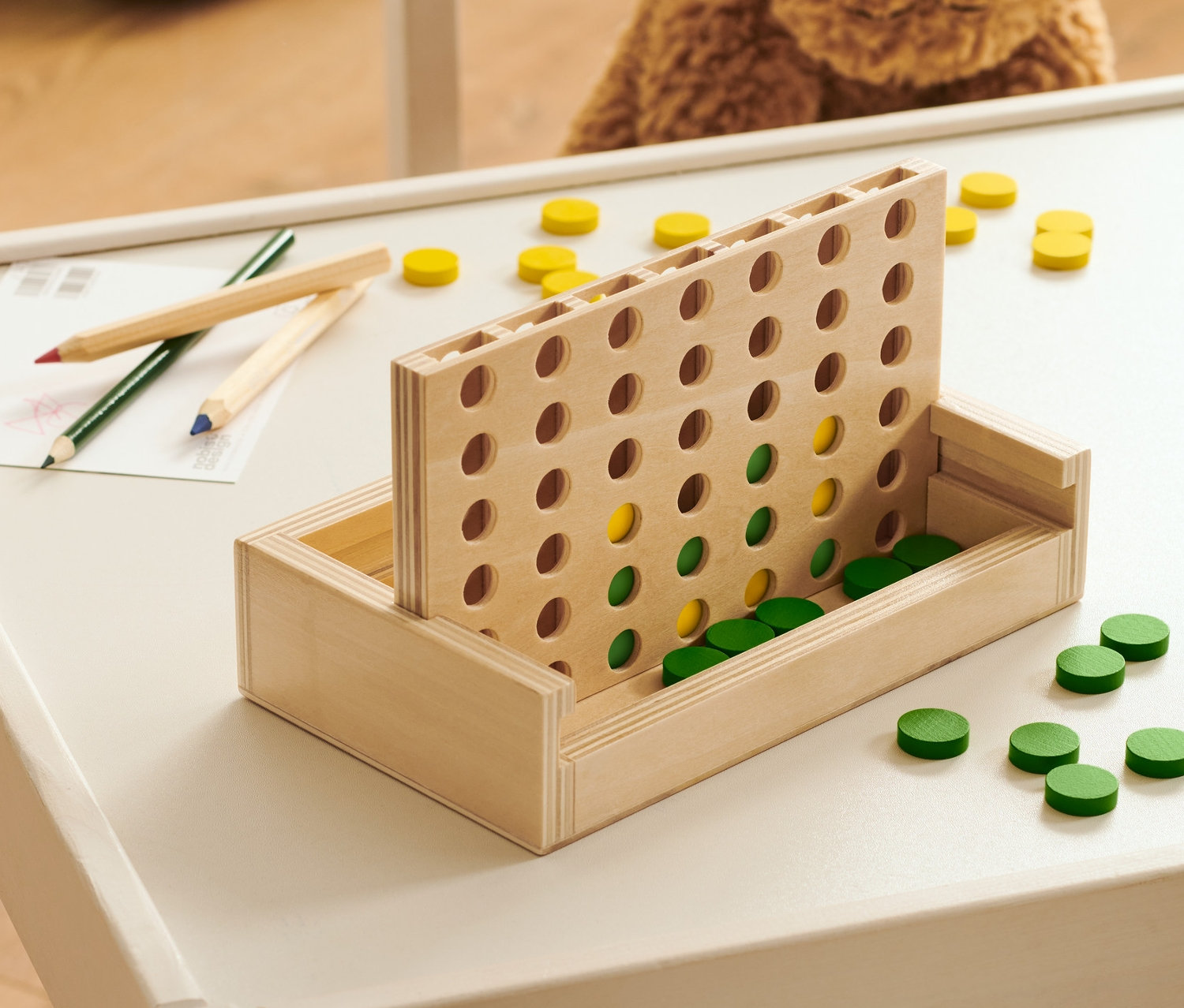 Holzspiel »4 in einer Reihe« steht mit Spielsteinen und Stiften auf einem Tisch.
