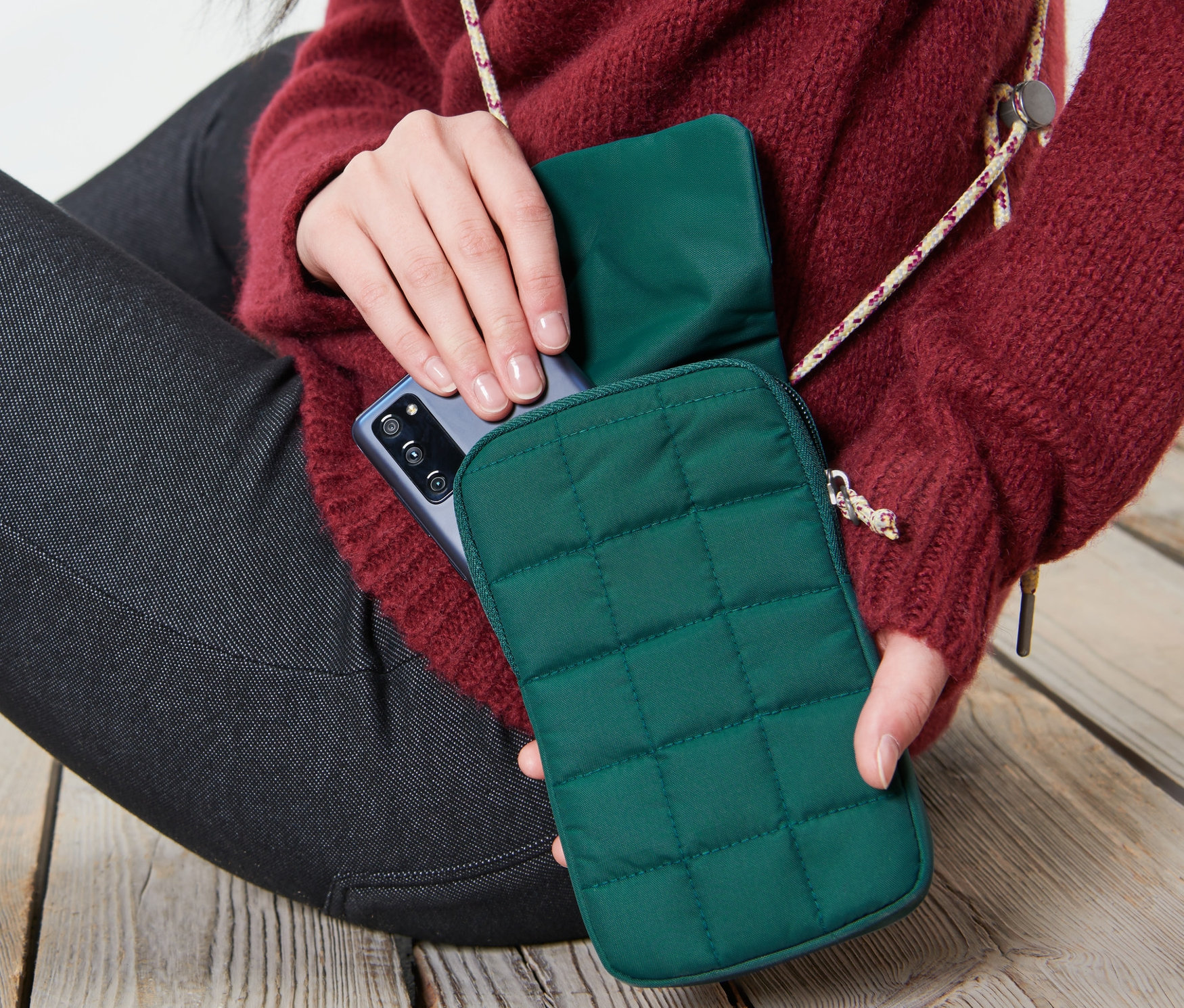 Frau sitzt auf Holzboden und legt ein Handy in eine dunkelgrüne isolierte Smartphone-Tasche.