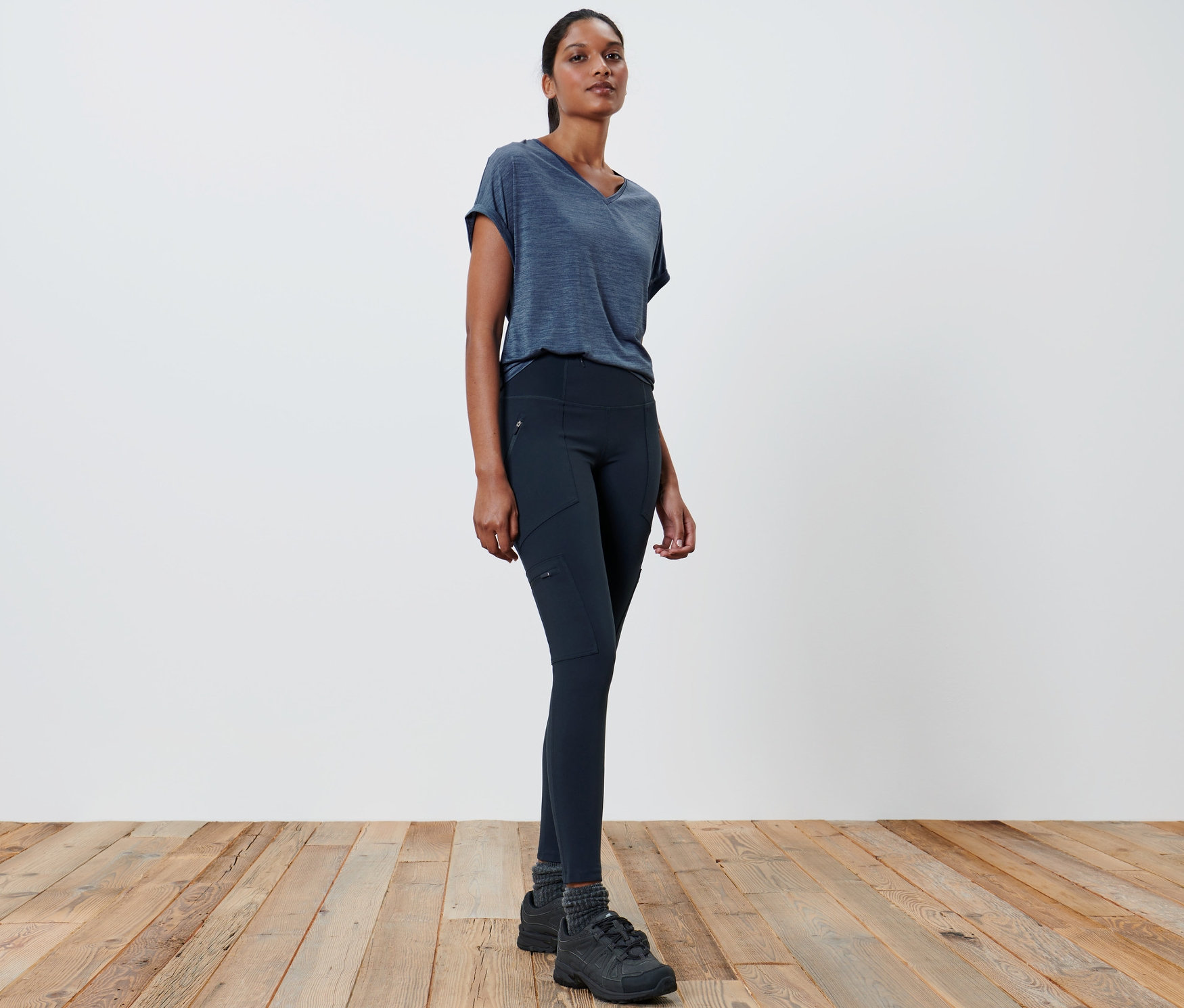 Frau in grauem T-Shirt und schwarzen Outdoor-Tight steht auf Holzboden.