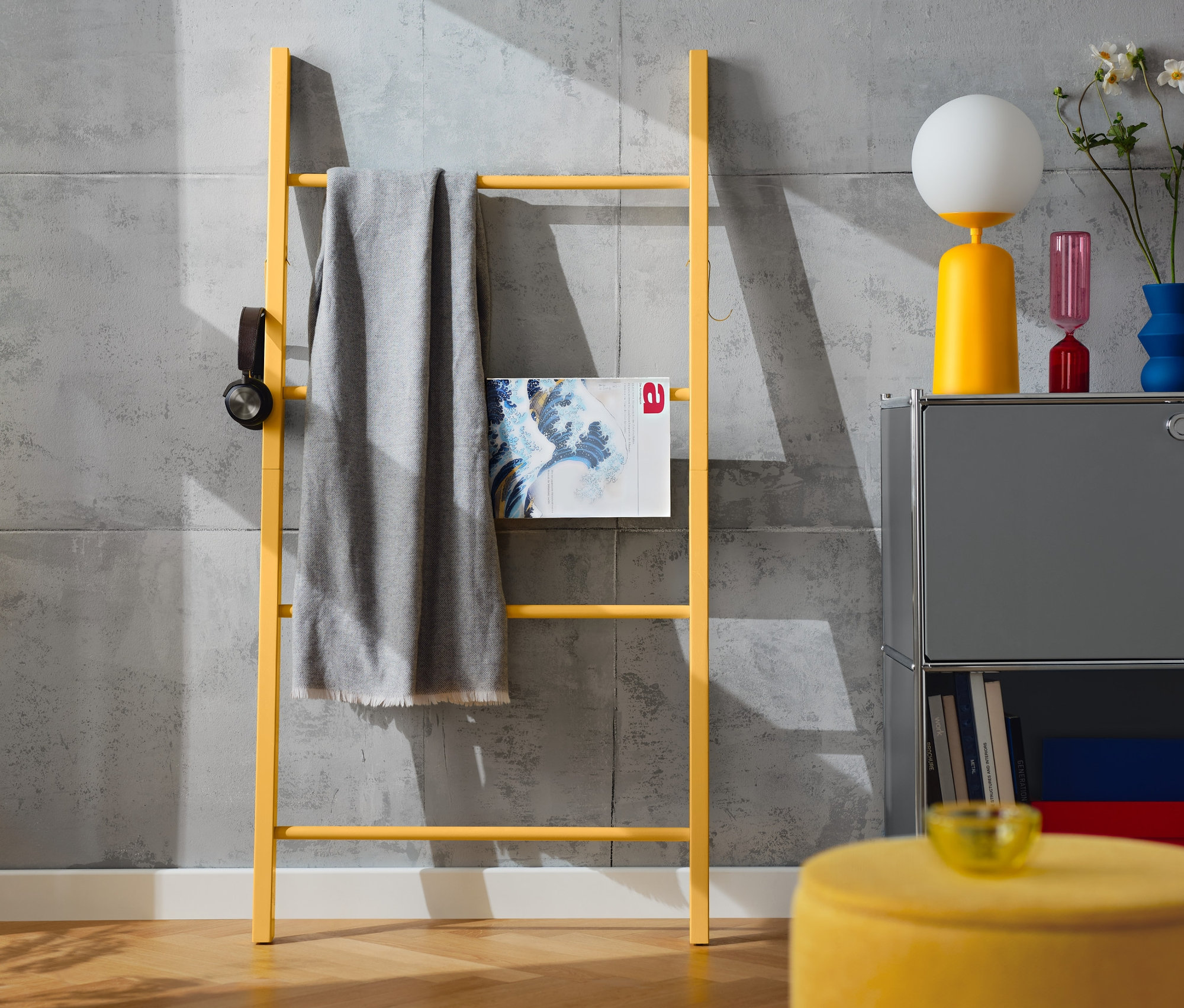 Eine gelbe Holzleiter mit Haken, an der eine graue Decke hängt. Daneben steht eine gelbe Tischleuchte mit Glaskugel.