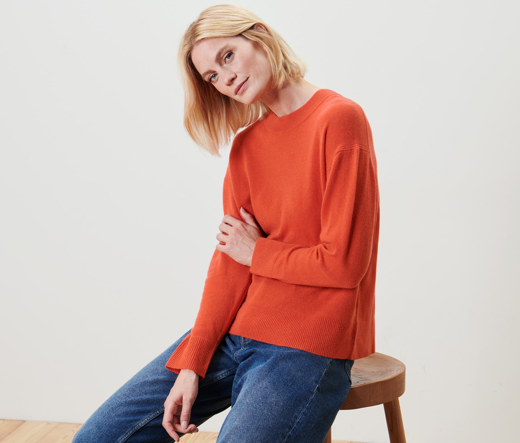 Blonde Frau sitzt auf einem Hocker und trägt einen orangefarbenen Feinstrick-Pullover mit Cashmere und Jeans.