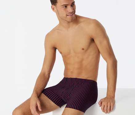 Ein Mann sitzt oberkörperfrei und blickt nach rechts, er trägt dunkelblaue SCHIESSER Shorts mit roten Streifen.