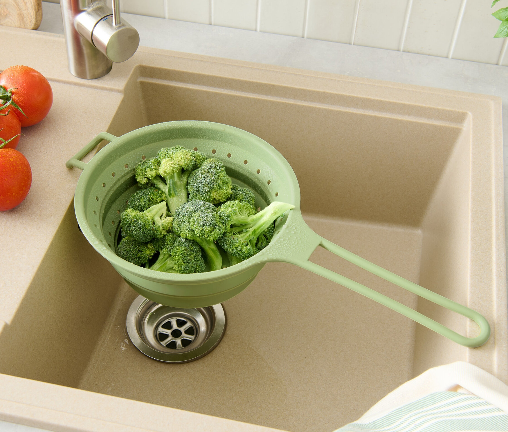 Brokkoli in einem grünen Platzspar-Sieb in einer beigen Spüle mit Tomaten und Kräutern.