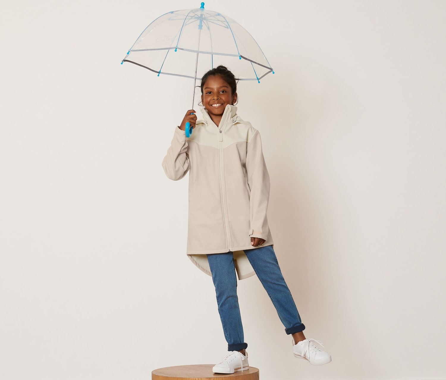 Ein Mädchen steht auf einem Holzsockel und hält einen durchsichtigen Regenschirm. Sie trägt einen Kinder-Softshellmantel.