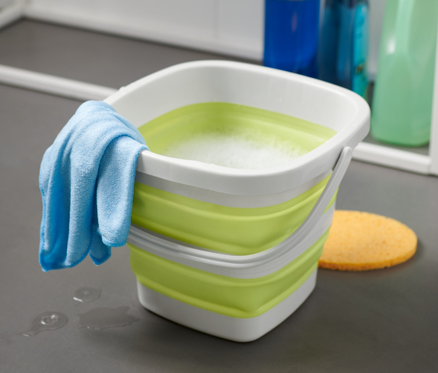 Ein faltbarer, eckiger Putzeimer mit einem Waschlappen und Schwamm auf dem Boden.